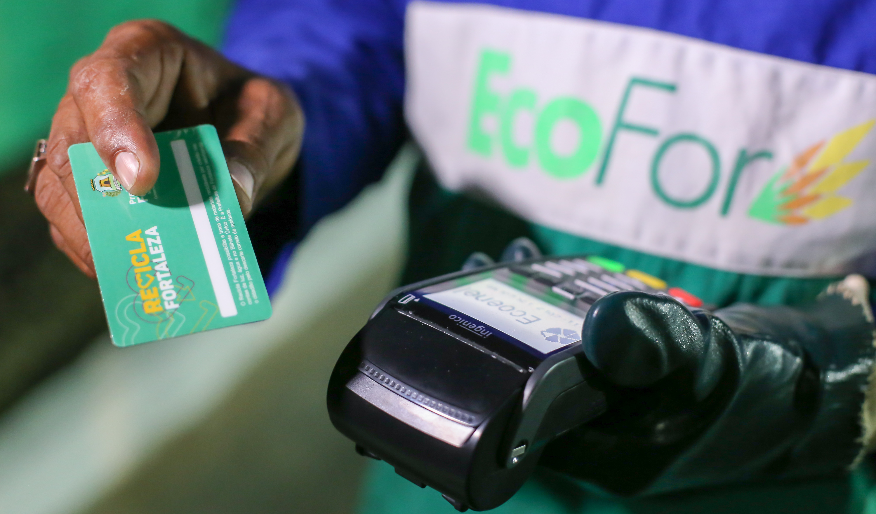 mão de funcionário vestido com farda da Ecofor Ambiental segurando cartão onde está escrito Recicla Fortaleza ao lado de máquineta de cartão